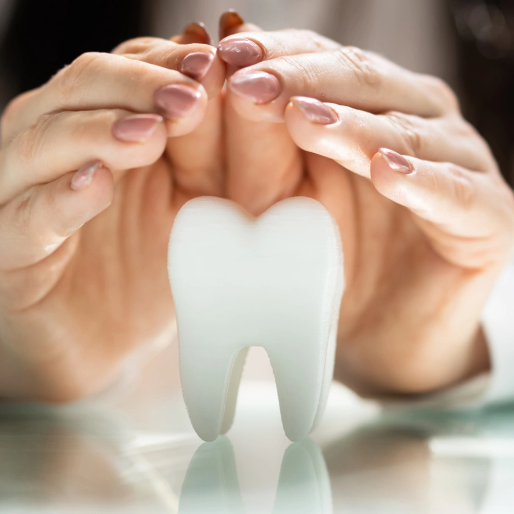 Hands forming heart shape above model tooth on dental office surface.